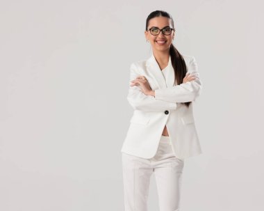 Gözlüklü, kollarını kavuşturup gri arka planda poz verirken gülümseyen beyaz takım elbiseli mutlu genç iş kadını.