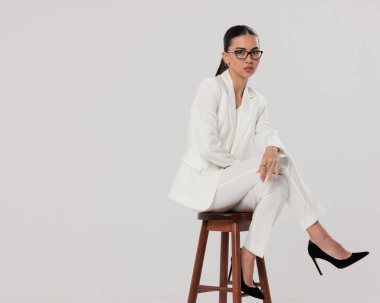 Gözlüklü, beyaz takım elbiseli, topuklu giyen ve dirseği gri arka planda oturan kendine güvenen bir iş kadını.