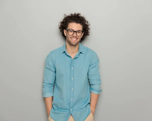 デニムシャツにメガネをかけた魅力的なカジュアルな男がポケットに手をつないで笑顔で前を向いて灰色の背景にポーズする — ストック写真