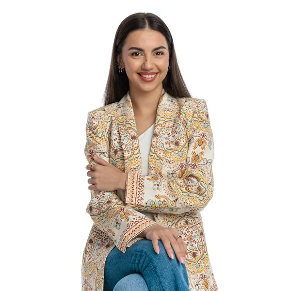 Çekici genç bir kadın ileriye bakıyor, kollarını kavuşturuyor ve avuçlarını dizlerinin üzerinde tutarken gülümsüyor ve beyaz arka planda poz veriyor.