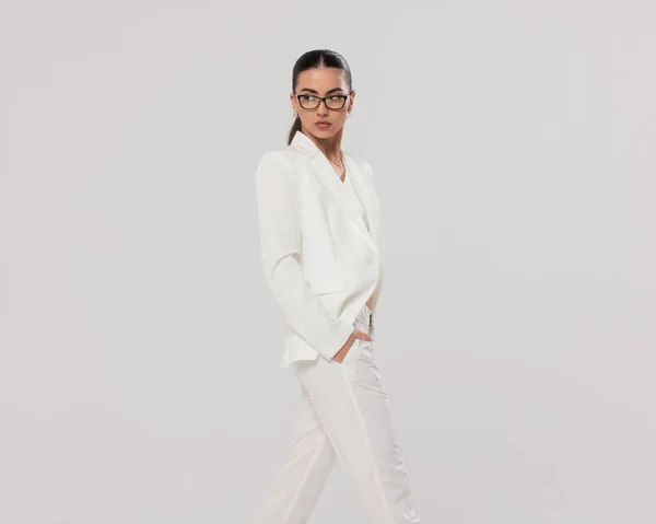 cool fashion woman with glasses in white suit holding hands in pockets and looking over shoulder while walking in front of grey background