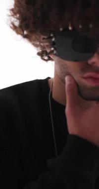 Yakışıklı, sıradan bir adam, çivili güneş gözlüklü, çenesine dokunan ve kibirli bir şekilde gri arka plan önünde yan yan yan bakan.