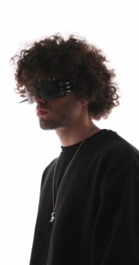 Kıvırcık saçlı, sivri uçlu güneş gözlüklü, ileriye bakan ve gri arka planda duran seksi adamın yan görüntüsü.