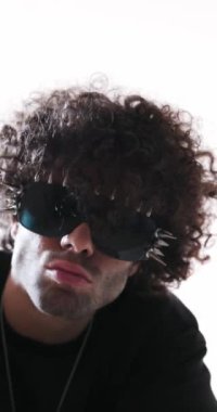 Siyah kapüşonlu, çivili güneş gözlüklü rahat bir adam kollarını kavuşturup gri arka planın önüne bakıyor.