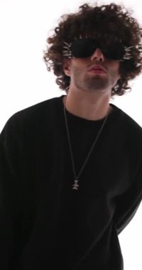 Siyah kazaklı yakışıklı, kıvırcık saçlı adam. Güneş gözlüğü takıyor, kollarını kavuşturuyor, çenesine dokunuyor ve gri arkaplan önünde düşünüyor.