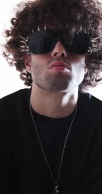 Sivri gözlükleri olan kibirli, ciddi bir adam kameraya yaklaşıyor, ileri bakıyor ve gri arka planda havalı davranıyor.