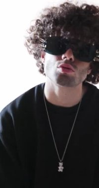 Kıvırcık saçlı, çivili güneş gözlüğü takan, sessiz bir jest yapan ve gri arka planda yürüyüp giden moda delikanlısı.