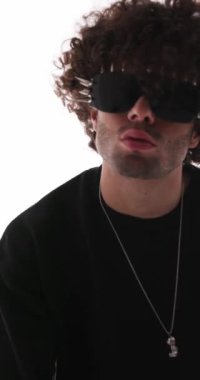 Kıvırcık saçlı, çivili güneş gözlüklü, sessiz hareketler yapan ileri ve geri dönüp gri arka planda ayrılan sıradan bir gencin dikey videosu.