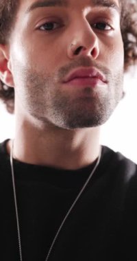 Siyah tişörtlü, kıvırcık saçlı, öne bakan, kollarını kavuşturan ve gri arka planda havalı bir şekilde gülümseyen yakışıklı adam.