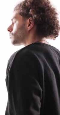 Havalı, kıvırcık saçlı, ileri bakan, hareket eden ve dönen moda adamı. Tişört yakasını düzeltirken ve kollarını gri arka planda kavuştururken.