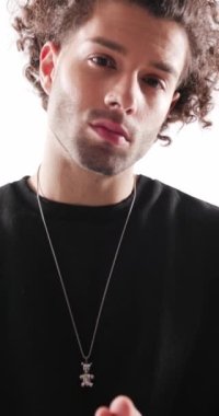 Kıvırcık saçlı, avuçlarını ovuşturan, ellerini boynuna koyan, dudaklarına dokunan, gri arka planda ileriye bakan seksi moda adamı.