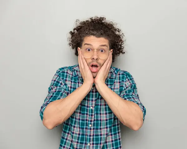 portrait of shocked man with glasses in plaid shirt opening mouth and being surprised while holding face in palms in front of grey background