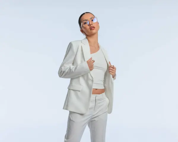 sexy elegant businesswoman in white suit with glasses adjusting suit while posing in a confident way in front of grey background
