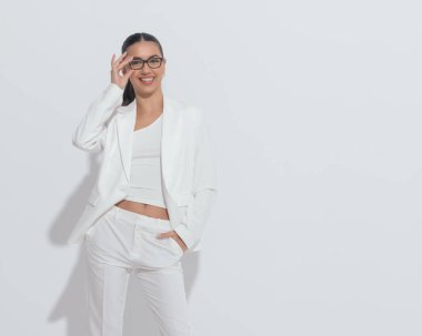 Zarif bir iş kadını gülüyor, güneş gözlüğünü tamir ediyor ve beyaz arka planda elleri ceplerinde poz veriyor. 