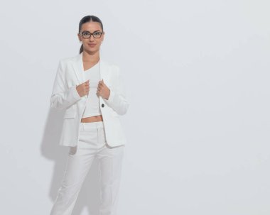 Gözlüklü, takım elbiseli ve gülümseyen güzel genç iş kadınının portresi. Gri arka planda durup poz verirken. 