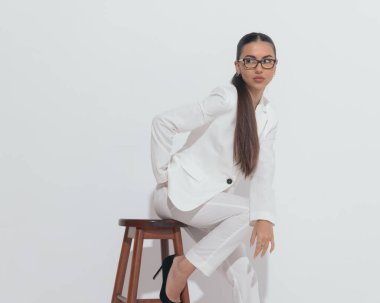 Gözlüklü, gözlüklü, el ele poz veren, kollarını dizlerinin üstüne koyup gri arka planda oturan zarif bir iş kadını.