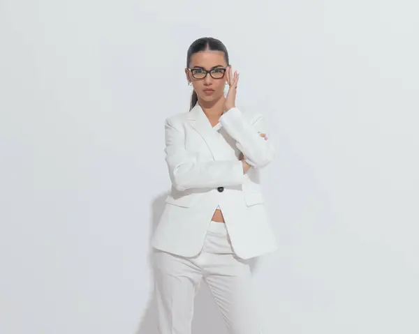 confident elegant woman folding arms and adjusting eyeglasses while standing and posing in front of grey background