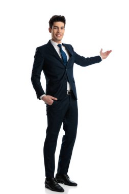 Mavi takım elbiseli, gururlu, zarif bir adamın tüm vücudu gülümseyen, bir elini cebinde beyaz arka planda tutarak sunan.