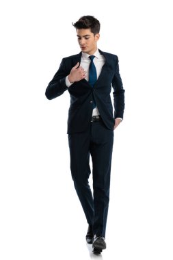 Seksi moda adamı el ele tutuşuyor, takım elbiseyi ayarlıyor ve beyaz arka planda yürürken aşağıya bakıyor.