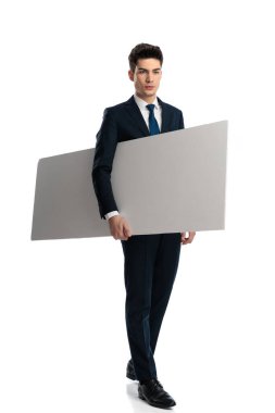 Lacivert takımlı seksi moda adamı kolunun altında boş ilan panosu tutuyor ve beyaz arka planda yürürken başka tarafa bakıyor.