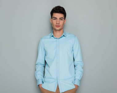 Mavi kot gömlekli çekici bir gencin portresi. Ceplerinde el ele tutuşup gri arka planda ileriye bakıyor.