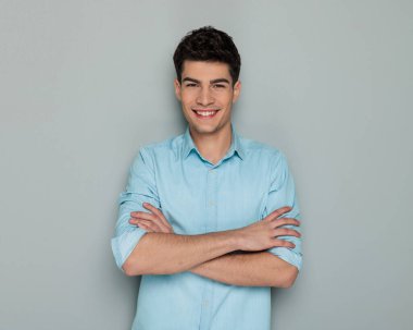 Kotlu gömlekli yakışıklı genç adam kollarını kavuşturuyor ve gri arka planda ileriye bakarken gülüyor.