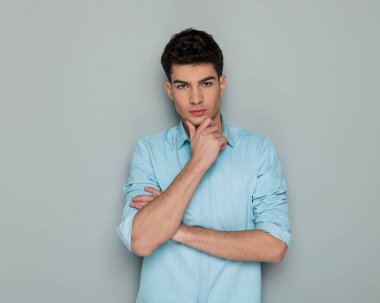 Mavi kot gömlekli, kollarını kavuşturan, çenesine dokunan ve gri arka planda düşünen kendinden emin genç bir adamın portresi.