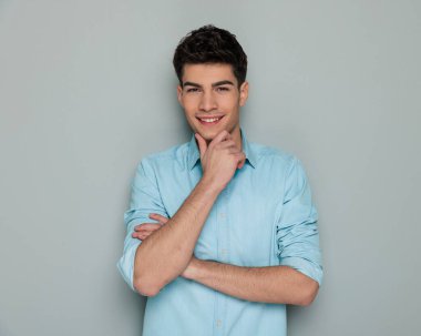 Mavi kot gömlekli mutlu adam kollarını katlıyor, çenesine dokunuyor ve gri arka planda hayal kuruyor.