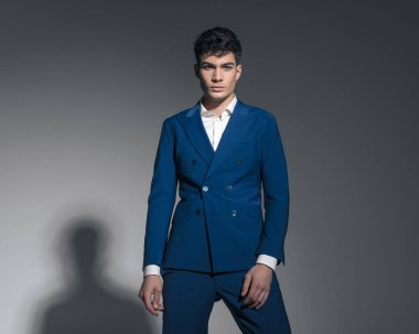 Açık gri arka planda dikilirken çift göğüslü mavi takım elbise giyen yakışıklı genç adamın portresi.