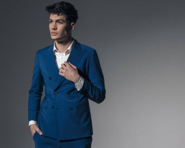 Gri arka planda çift göğüslü mavi yakasını düzeltirken cebi tutan yakışıklı bir genç.