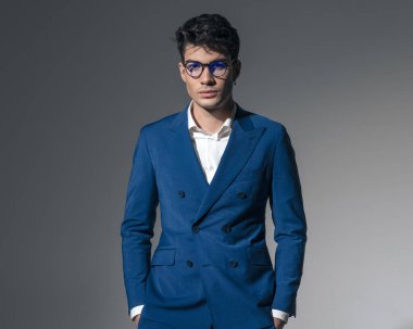 Açık gri arka planda elleri ceplerinde gözlük takan rahat ve zarif bir adamın portresi.