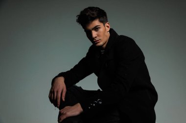 Gri arka planda havalı bir şekilde poz verirken siyah ceketli, dizinin üstünde oturan ve bacağını tutan çekici genç adamın yan görüntüsü.