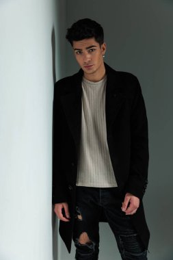 Siyah ceketli çekici bir adam duvara yaslanıyor ve gri arka planda gizemli bir şekilde poz veriyor.