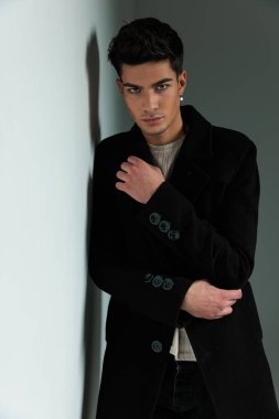 Siyah ceketli dramatik havalı adamın portresi. Duvara yaslanıyor, kollarını kavuşturuyor ve gri arka planda ileri doğru bakıyor.