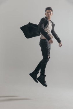 Genç adam havada paltoyla zıplıyor ve gri arka planda yer almak istiyor.