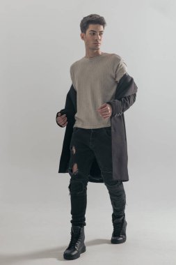 Gri arka planda siyah paltosunu çıkarırken ileri doğru yürüyen meraklı genç adam.