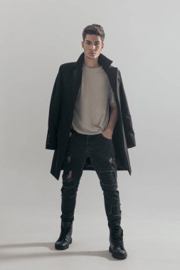 Omuzlarına palto giymiş rahat bir adam elleri ceplerinde açık gri arka planda dikiliyor.