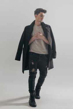 Siyah ceketli çekici genç adam açık gri arka planda yürürken el ele tutuşuyor ve aşağıya bakıyor.