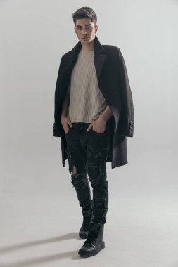 Siyah paltolu rahat genç adam elleri ceplerinde gri arka planda duruyor.