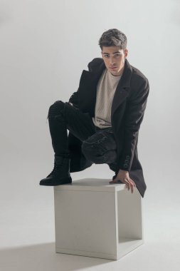 Siyah paltolu yakışıklı bir adam sandalyede duruyor ve gri arka plana yaslanıyor.