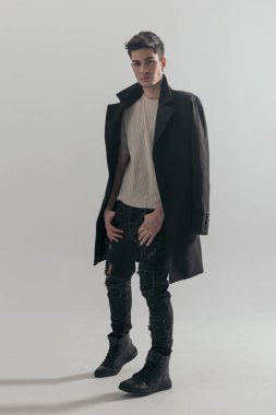 Açık gri arka planda siyah palto giyerken cebi tutan seksi, sıradan bir adam.