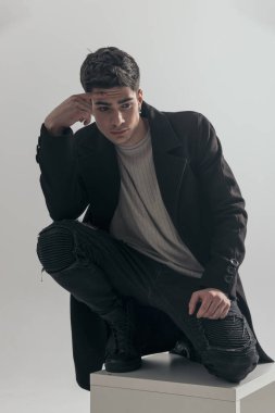 Düşünceli genç adam sandalyede çömelmiş açık gri arka planda aşağı doğru bakıyor.