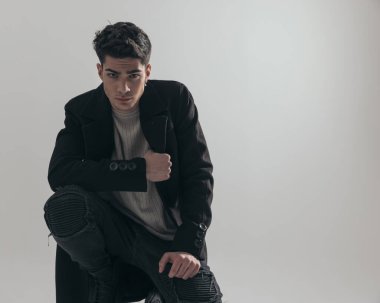 Gri arka planda çömelmiş siyah paltolu çekici, sıradan bir adama yakın çekim.