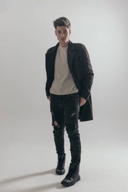 Gri arka planda yürüyen, siyah paltolu ve kot pantolonlu rahat bir adam.