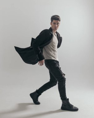 Seksi bir adam gri arka planda yürüyor. Havada siyah bir palto var. Uzun bir resim.