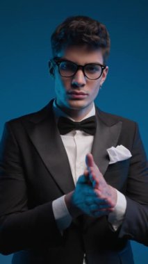Gözlüklü, zarif, siyah smokinli, avuçlarını ovuşturan ve renkli arka planda ileriye bakan çekici bir gencin video projesi.