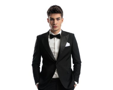 Siyah smokinli çekici bir adamın portresi. Ceplerinde el ele tutuşup beyaz arka planda poz verirken ileriye bakıyor.