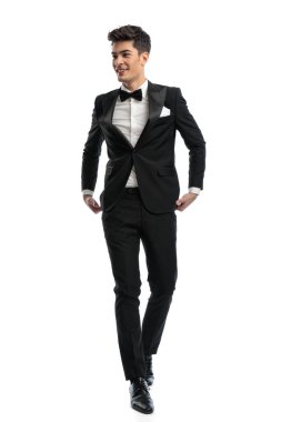 Mutlu zarif iş adamı beyaz arka planda yürürken gülümseyip smokin ayarlıyor.