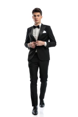 Beyaz arkaplan önünde siyah smokini kapatırken ileri ve geri yürüyen kendine güvenen zarif iş adamının uzun bir resmi. 