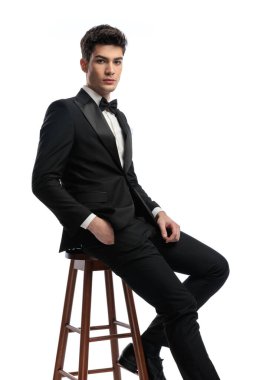 Siyah smokinli kendine güvenen genç damat cebinde oturuyor ve beyaz arka planda ileriye bakıyor.